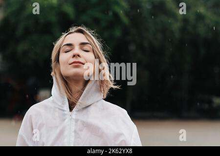 Gros plan Portrait de la jeune femme portant un imperméable en se tenant sur la rue à Rainy Day, Blonde fille vêtue de manteau imperméable ayant du plaisir sous Banque D'Images