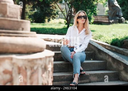 Élégante femme caucasienne assise sur les escaliers dans le parc public et tenant le smartphone à la main, confiante femme Entrepreneur portant des lunettes de soleil et Banque D'Images