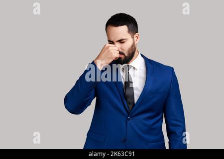 Portrait d'un homme barbu grimaçant avec dégoût, tenant l'haleine, pinçant le nez avec les doigts pour éviter la mauvaise odeur, odeur horrible, portant un costume de style officiel. Prise de vue en studio isolée sur fond gris. Banque D'Images