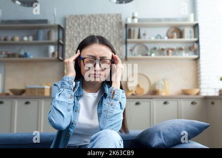 Une jeune femme asiatique malade est assise sur le canapé à la maison, la femme a un mal de tête, une photo rapprochée d'un étudiant en lunettes. Banque D'Images