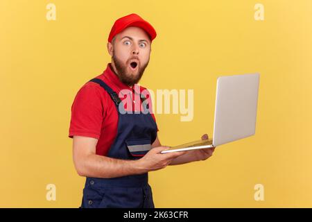 Vue latérale d'un magnifique homme de main surpris debout dans les mains et travaillant en ligne, regardant la caméra avec les yeux grands, portant uniforme et capuchon rouge. Studio d'intérieur isolé sur fond jaune. Banque D'Images