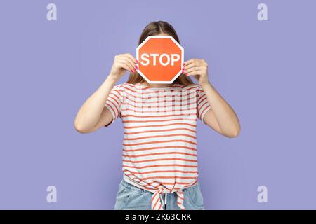 Portrait d'une femme inconnue portant un T-shirt tenant et cachant son visage avec un panneau d'arrêt rouge, tenant un panneau de signalisation rouge, l'attention au passage à niveau sécuritaire. Studio d'intérieur isolé sur fond violet. Banque D'Images