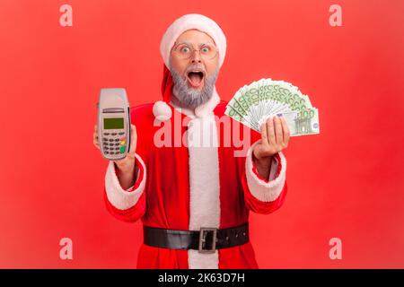 Homme âgé étonné avec une barbe grise portant le costume du père noël debout à bouche ouverte, tenant le terminal et les billets en euros entre les mains. Studio d'intérieur isolé sur fond rouge. Banque D'Images