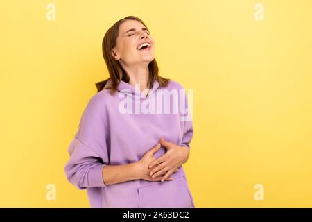 Portrait d'une femme heureuse avec des cheveux foncés tenant le ventre, riant, entendant une blague ou une anecdote drôle, étant de bonne humeur, portant le sweat à capuche violet. Studio d'intérieur isolé sur fond jaune. Banque D'Images