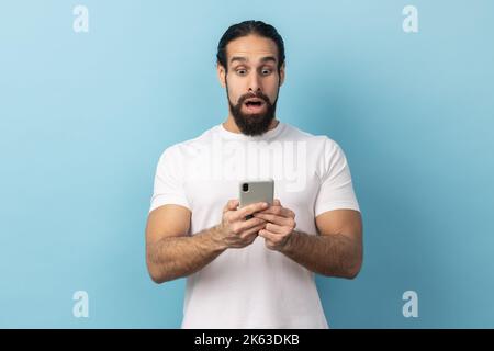 Portrait d'un homme surpris avec une barbe portant un T-shirt blanc exprimant un choc lors de l'utilisation d'un téléphone portable, la recherche sur le Web, des nouvelles étonnantes. Studio d'intérieur isolé sur fond bleu. Banque D'Images