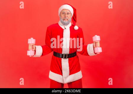 Portrait d'un homme fort âgé avec une barbe grise portant le costume du père noël debout avec des haltères entre les mains, essayant de soulever des équipements de sport lourds. Studio d'intérieur isolé sur fond rouge. Banque D'Images