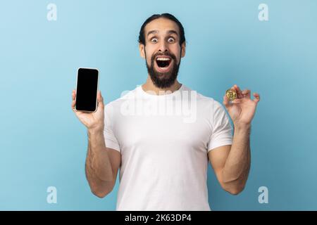 Portrait d'un homme émerveillé avec une barbe portant un T-shirt blanc tenant un téléphone portable et un bitcoin doré, publicité des applications crypto. Studio d'intérieur isolé sur fond bleu. Banque D'Images