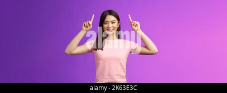 Adorable femme asiatique aux cheveux sombres et affirmée porter un t-shirt rose lever les mains, montrer la promotion, pointer les doigts vers le haut, sourire, inviter utiliser le lien, essayez Banque D'Images