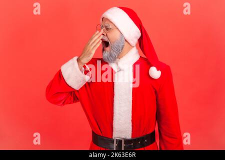 Homme âgé avec barbe grise portant le costume du père noël bâchant et couvrant la bouche avec le bras, se sentant somnolent après une nuit sans sommeil, manque d'énergie. Studio d'intérieur isolé sur fond rouge. Banque D'Images