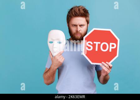 Portrait d'un homme barbu sérieux et bossy tenant un panneau d'arrêt rouge et un masque blanc, regardant un appareil photo avec une expression stricte. Studio d'intérieur isolé sur fond bleu. Banque D'Images