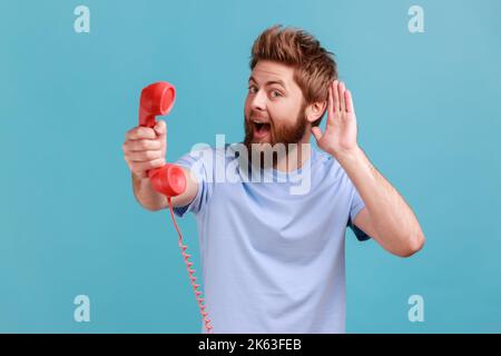 Portrait d'un homme barbu tenant et montrant le combiné rétro du téléphone à l'appareil photo, demandant de répondre au téléphone, garde la main près de l'oreille, entendant votre conversation. Studio d'intérieur isolé sur fond bleu. Banque D'Images