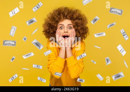 Oh mon dieu, wow. Portrait d'une femme avec une coiffure afro regardant l'appareil photo avec de grands yeux, absolument choqué de la pluie d'argent tombant de haut. Studio d'intérieur isolé sur fond jaune. Banque D'Images