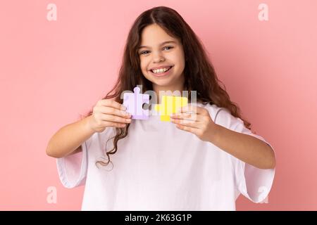 Portrait d'une petite fille souriante et satisfaite portant un T-shirt blanc tenant des pièces de puzzle jaunes et violets, résoudre la tâche, regarder l'appareil photo. Studio d'intérieur isolé sur fond rose. Banque D'Images