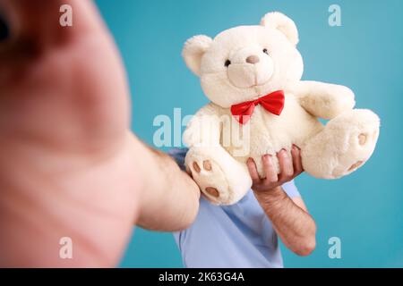Portrait d'un homme barbu inconnu qui fait du selfie ou diffuse en direct, point de vue photo, cachant son visage derrière un ours en peluche. Studio d'intérieur isolé sur fond bleu. Banque D'Images