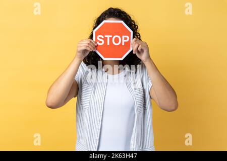 Anonyme femme avec des cheveux ondulés foncé debout couvrant son visage avec le signe d'arrêt rouge, avertissement sur les problèmes, concept de l'interdiction, déni, interdit d'aller. Studio d'intérieur isolé sur fond jaune. Banque D'Images