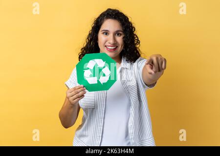 Portrait d'une femme souriante satisfaite ravie avec des cheveux ondulés foncé tenant le panneau vert de recyclage et pointant vers l'appareil photo, en économisant l'environnement. Studio d'intérieur isolé sur fond jaune. Banque D'Images