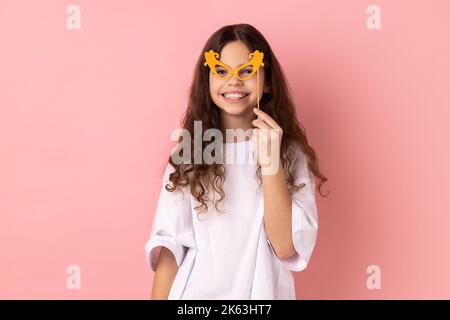 Portrait d'une jolie petite fille heureuse portant un T-shirt blanc souriant et tenant des verres en papier sur le bâton, en utilisant des accessoires de fête en vacances, en s'amusant. Studio d'intérieur isolé sur fond rose. Banque D'Images