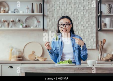 Portrait d'une femme heureuse à la maison, femme asiatique mangeant une salade de nourriture saine et souriante, femme au foyer regardant l'appareil photo assis dans la cuisine à table, régime pour la perte de poids et la forme physique. Banque D'Images