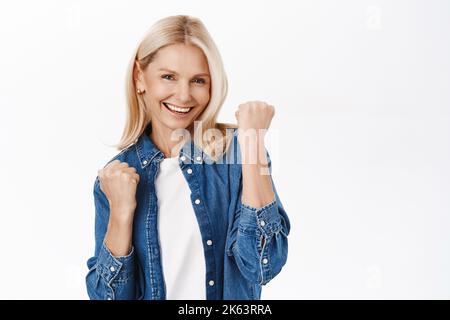 Oui, réussite. Femme mûre blonde excitée célébrant, rend la pompe de poing et les sourires satisfaits, atteindre le but, triomphent, se dresse sur fond blanc Banque D'Images