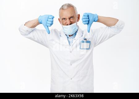 Coronavirus et concept de santé. Médecin en colère dans le masque médical, montrant les pouces vers le bas geste détestant, disapprouver smth, debout sur blanc Banque D'Images