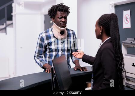 Homme avec collier de cou au comptoir de réception, demandant de l'aide et d'être dans la douleur. Patient blessé avec de la mousse cervicale attendant d'assister à un rendez-vous d'examen de contrôle pour guérir la blessure après accident. Banque D'Images