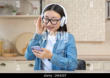 Jeune femme asiatique à la maison utilisant des écouteurs et un téléphone mobile pour écouter des podcasts en ligne, de la musique et des livres audio, femme en gros plan dans des lunettes dans la cuisine souriante et heureuse. Banque D'Images