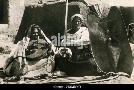 Couple Touareg algérien - Femme jouant un Izmad, un violon à corde unique joué uniquement par des femmes Touareg. L'instrument unique est fabriqué à partir d'une demi-coquille de gourde recouverte de peau d'animal, d'un manche en bois et d'une ficelle de poils de cheval. Les hommes risquent une malédiction s'ils jouent de l'instrument, selon une croyance ancienne. Le monsieur avec l'épée fine et le bouclier est plutôt nain dans les enjeux d'intérêt ayant considéré l'Izmad.... Banque D'Images