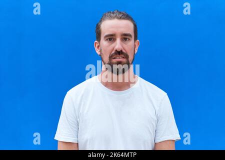 Jeune homme beau portant un t-shirt décontracté debout sur fond bleu isolé avec une expression sérieuse sur le visage. Banque D'Images