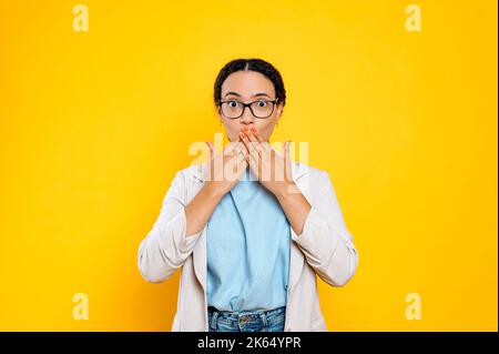 Excitée stupéfait latino ou brésilienne jeune femme d'affaires avec des lunettes, couvrant la bouche avec les mains, ne veut pas trop dire, regarde la caméra, se tient sur un arrière-plan isolé orange Banque D'Images