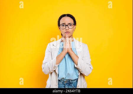 Demander quelque chose. Pleuding mignon brésilien ou latino jeune brune femme, employé de compagnie, tient les mains dans un panneau plié dans la prière, regarde la caméra, se tient sur un arrière-plan isolé orange Banque D'Images