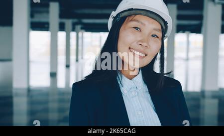 Portrait d'une femme asiatique ingénieur debout à l'intérieur d'un nouveau bâtiment portant un casque de sécurité souriant appréciant le travail. Concept de personnes et de professions. Banque D'Images