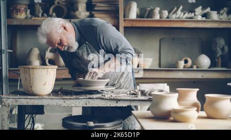 Un maître expérimenté est en train de faire un bol d'argile bas sur une roue de poterie dans un petit atelier de maison. Le puzzle en céramique, les articles de clayware faits à la main et les outils de potter sont visibles. Banque D'Images