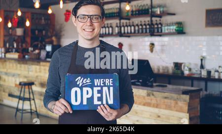 Portrait of cheerful handsome young waiter in apron holding we are open sign standing in opening coffee shop. Starting new business and successful people concept. Banque D'Images