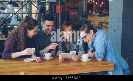 Les étudiants heureux et de bonne apparence regardent une vidéo amusante, en discutent et rient pendant la réunion dans un café. Des gens joyeux, une technologie moderne et un concept de restauration. Banque D'Images