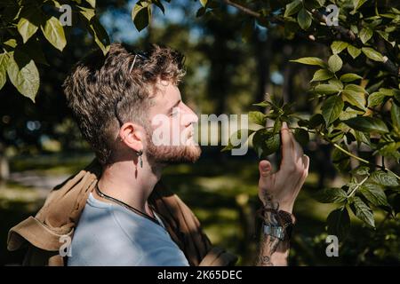 Un portrait d'un jeune homme blond qui sent des feuilles vertes dans un parc au printemps en plein soleil Banque D'Images