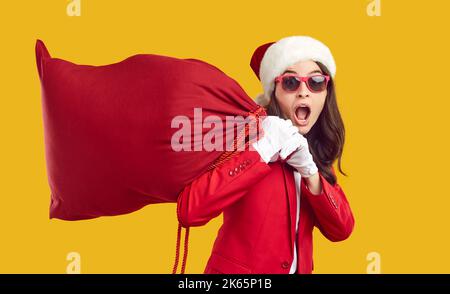 Drôle surprise femme en chapeau de Santa et lunettes de soleil portant sac rouge avec des cadeaux de Noël Banque D'Images