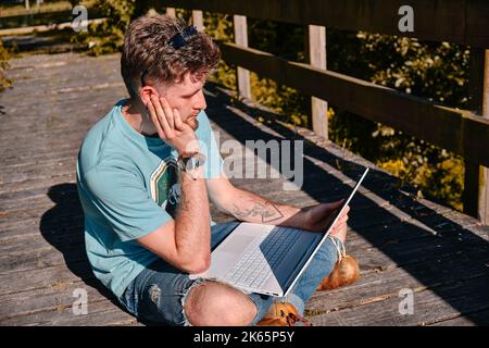 Un entrepreneur frustré travaillant avec un ordinateur portable sur un pont en bois dans un parc au soleil Banque D'Images