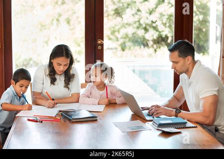 Jeune mère de race mixte aidant ses enfants à faire leurs devoirs pendant que leur père travaille sur un ordinateur portable à une table dans le salon. Dessin de petits frères et sœurs Banque D'Images