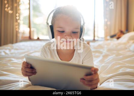 Petite fille avec une tablette numérique et un casque sur le lit à la maison. Un enfant mignon qui se fait un plaisir de regarder des dessins animés avec de la musique sur son appareil ou de faire un Banque D'Images