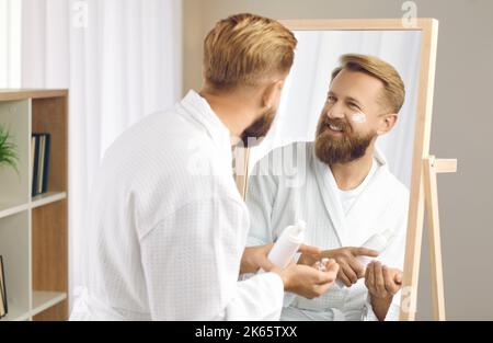 Homme heureux regardant son visage dans le miroir tout en faisant sa routine de soin de peau du matin Banque D'Images