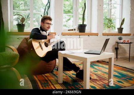 Jeune homme qui simord sur le sol à la maison et qui se penche pour jouer de la guitare. Passe-temps et activités de loisirs. Formation à distance, cours en ligne Banque D'Images