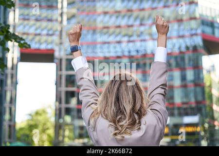 Professionnels. Vue arrière d'une femme d'affaires qui se tient debout dans la rue et qui tient la main dans la fête, triomphant, atteignant son but et se réjouissant Banque D'Images