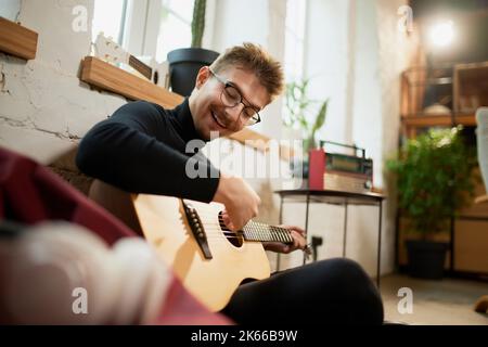 Jeune homme qui simord sur le sol à la maison et qui se penche pour jouer de la guitare. Passe-temps et activités de loisirs. Formation à distance, cours en ligne Banque D'Images