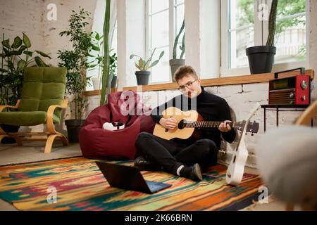 Jeune homme qui simord sur le sol à la maison et qui se penche pour jouer de la guitare. Passe-temps et activités de loisirs. Formation à distance, cours en ligne Banque D'Images