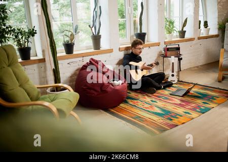 Jeune homme qui simord sur le sol à la maison et qui se penche pour jouer de la guitare. Passe-temps et activités de loisirs. Formation à distance, cours en ligne Banque D'Images