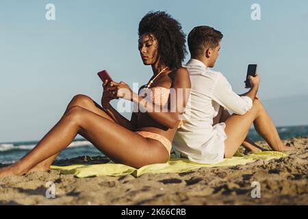 Un jeune couple multiethnique qui se couche d'épaule à épaule sur la plage en été - les jeunes sont toujours connectés à Internet - la technologie Banque D'Images