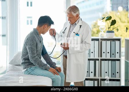 Un homme de race blanche mature médecin effectuant un examen de contrôle de la tension artérielle d'un jeune patient à l'hôpital. Un jeune homme asiatique assis sur un lit fait la routine Banque D'Images
