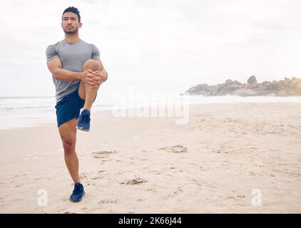 Passez à une séance d'entraînement sur la plage. Un jeune homme sportif s'étirant les jambes tout en s'exerçant sur la plage. Banque D'Images