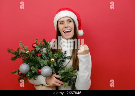 Gaie jeune femme caucasienne dans le chapeau du Père Noël tenant le bouquet de branches d'épinette faisant le geste gagnant célébrer les poings de clenching dire oui, posant est Banque D'Images