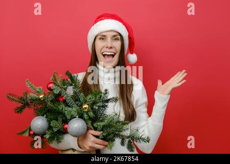 La jeune fille de père Noël choquée 20s porte un chapeau de Noël tenant un bouquet de branches d'épinette main levée, posant isolé sur le mur de fond de couleur rouge dans le goujon Banque D'Images